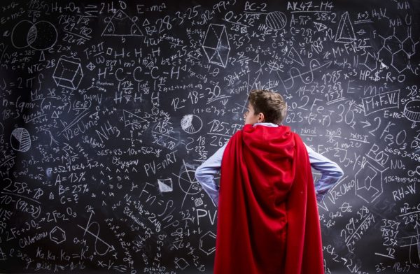 Młody mężczyzna w pelerynie bohatera stoi przodem do tablicy zapisanej równaniami
