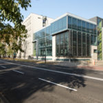 Przeszklony budynek z perspektywy drugiej strony ulicy, na zbliżeniu widać ruchliwą drogą i samochody